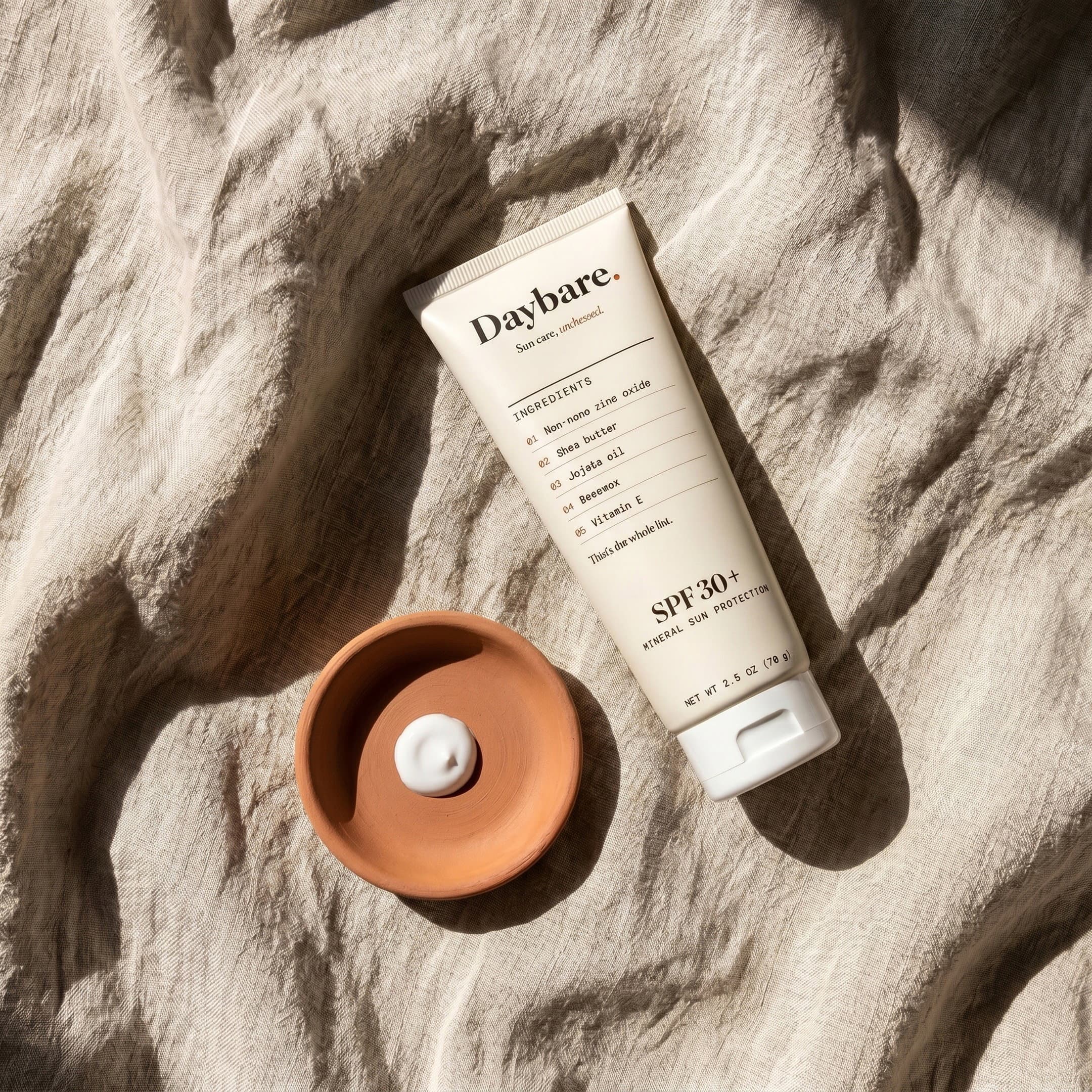 Daybare tube on raw linen beside a terracotta dish holding one smear of zinc balm